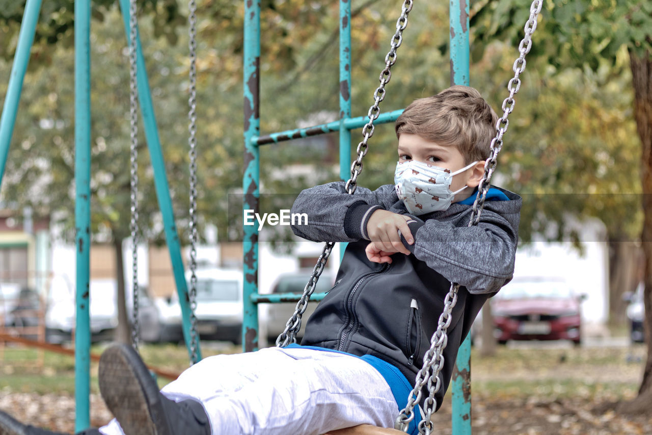Little boy wearing protective face mask while swinging in the park and looking at camera.