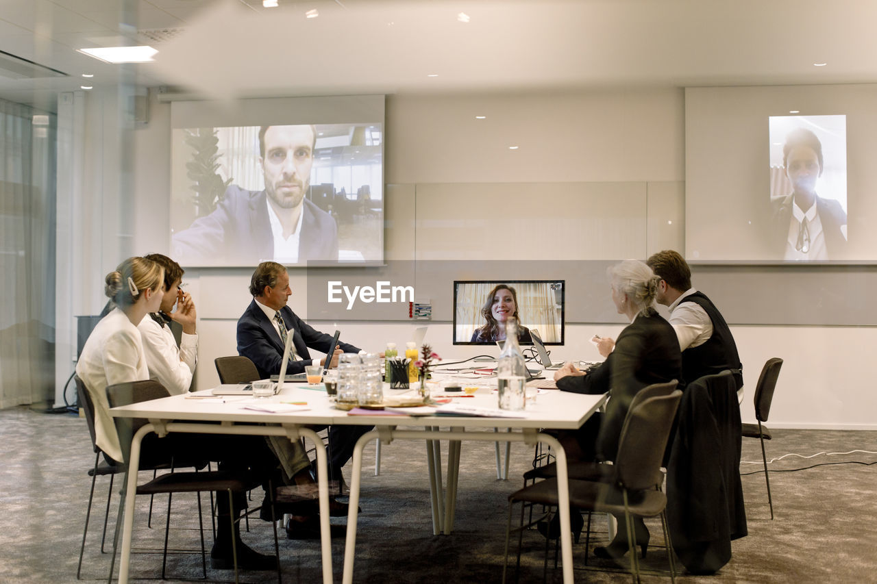 Male and female professionals discussing in global business meeting seen through glass wall at workplace