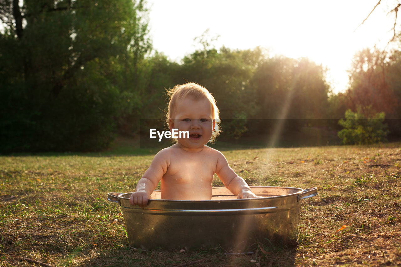 Cute little kid sits in a basin of water in nature and has rays of the setting sun, golden bathe. 