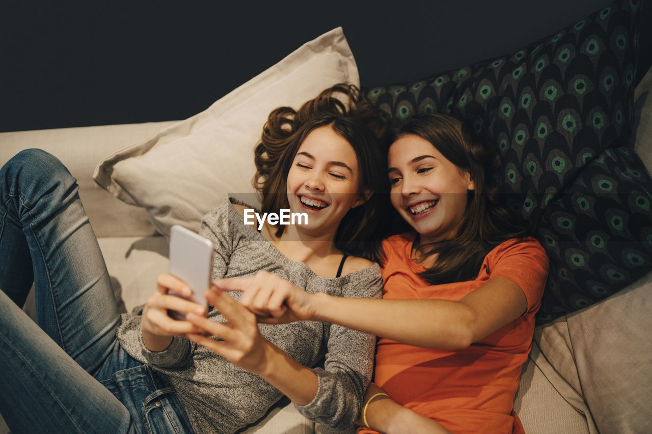High angle view of happy friends using phone while resting on sofa at home