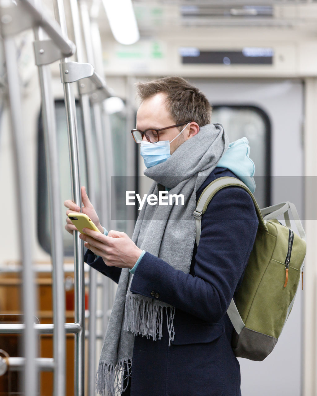 Side view of man wearing mask using smart phone while standing in subway train