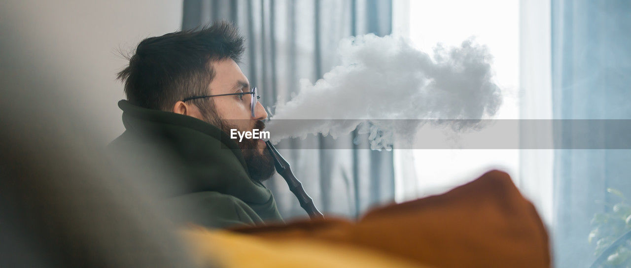 one person, adult, indoors, smoke, window, communication, men, blue, glasses, eyeglasses, business, businessman, young adult, looking, headshot, office, day, selective focus, portrait, side view, nature, activity, technology, contemplation, occupation, sitting, cloud, furniture