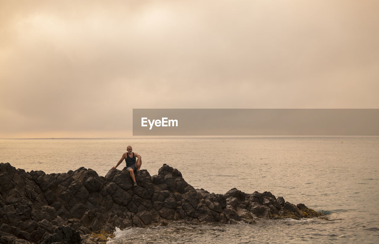 Man sitting on rock looking at scenic view of beach against sky