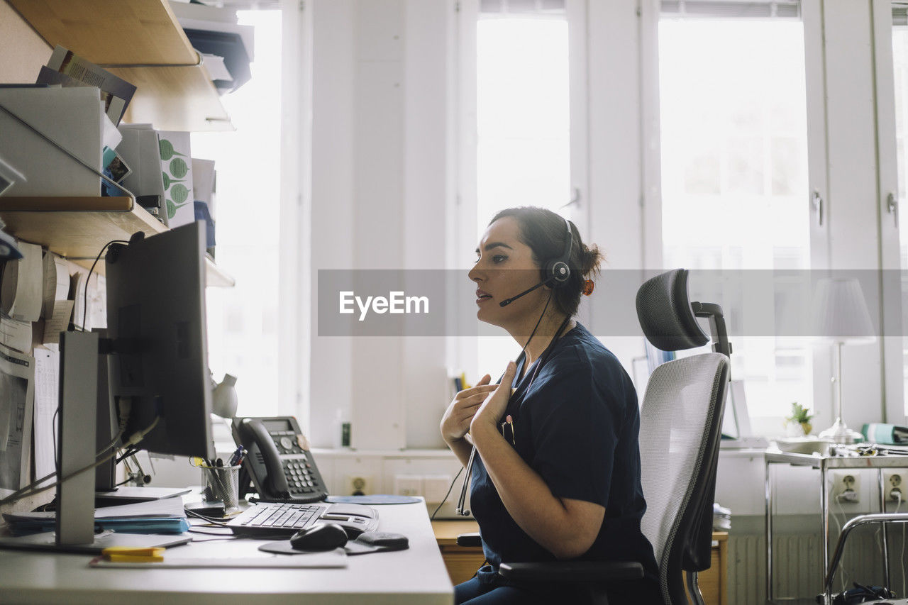 Side view of mature female nurse doing video call while sitting on chair in clinic