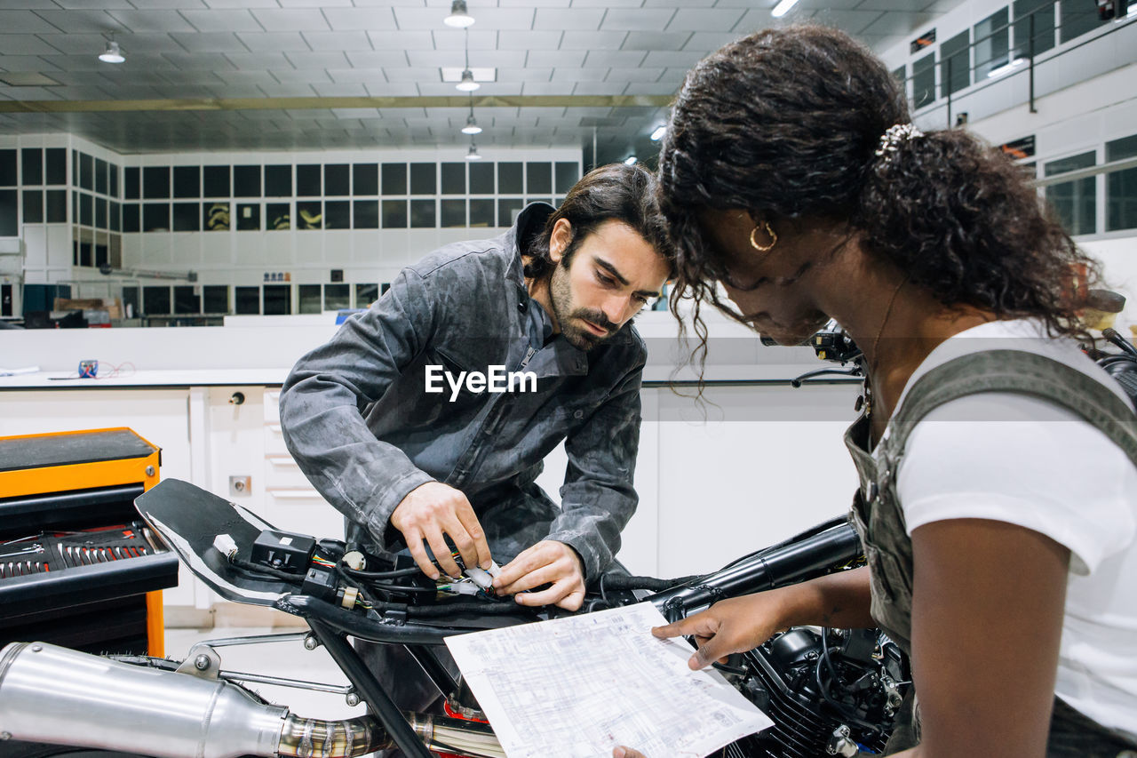 Side view of multiracial male and female mechanics repairing custom motorbike in spacious workshop while working together