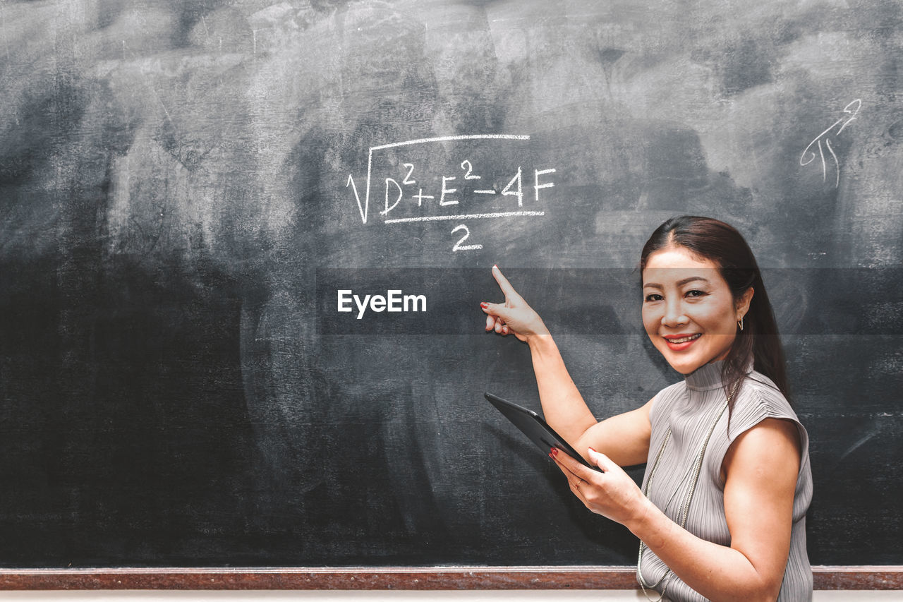 Portrait of teacher pointing at formula on blackboard in classroom
