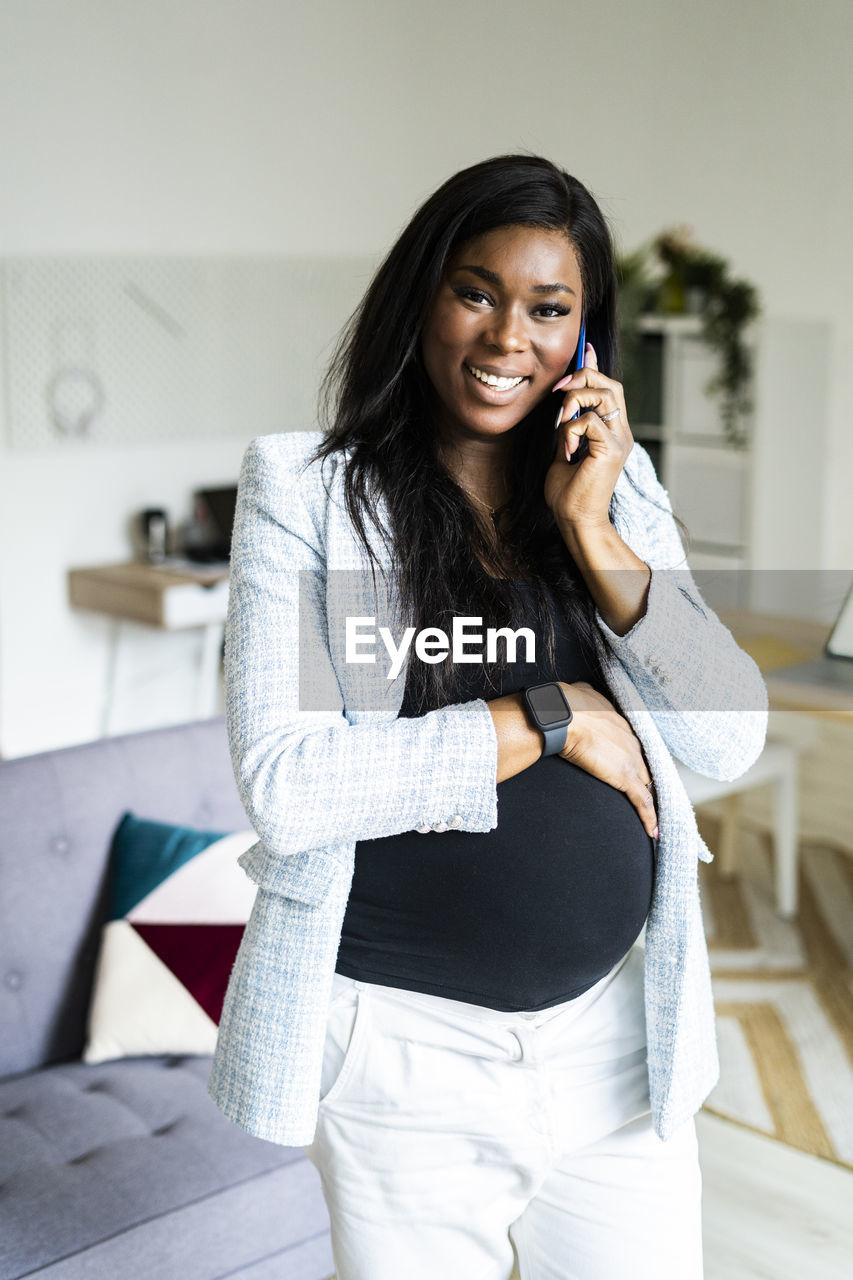 Happy pregnant businesswoman talking on smart phone while standing with hand on stomach