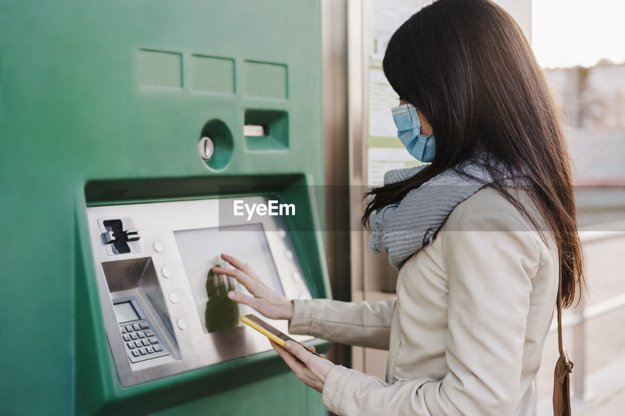 Woman wearing protective face mask operating ticket machine while holding mobile phone during pandemic