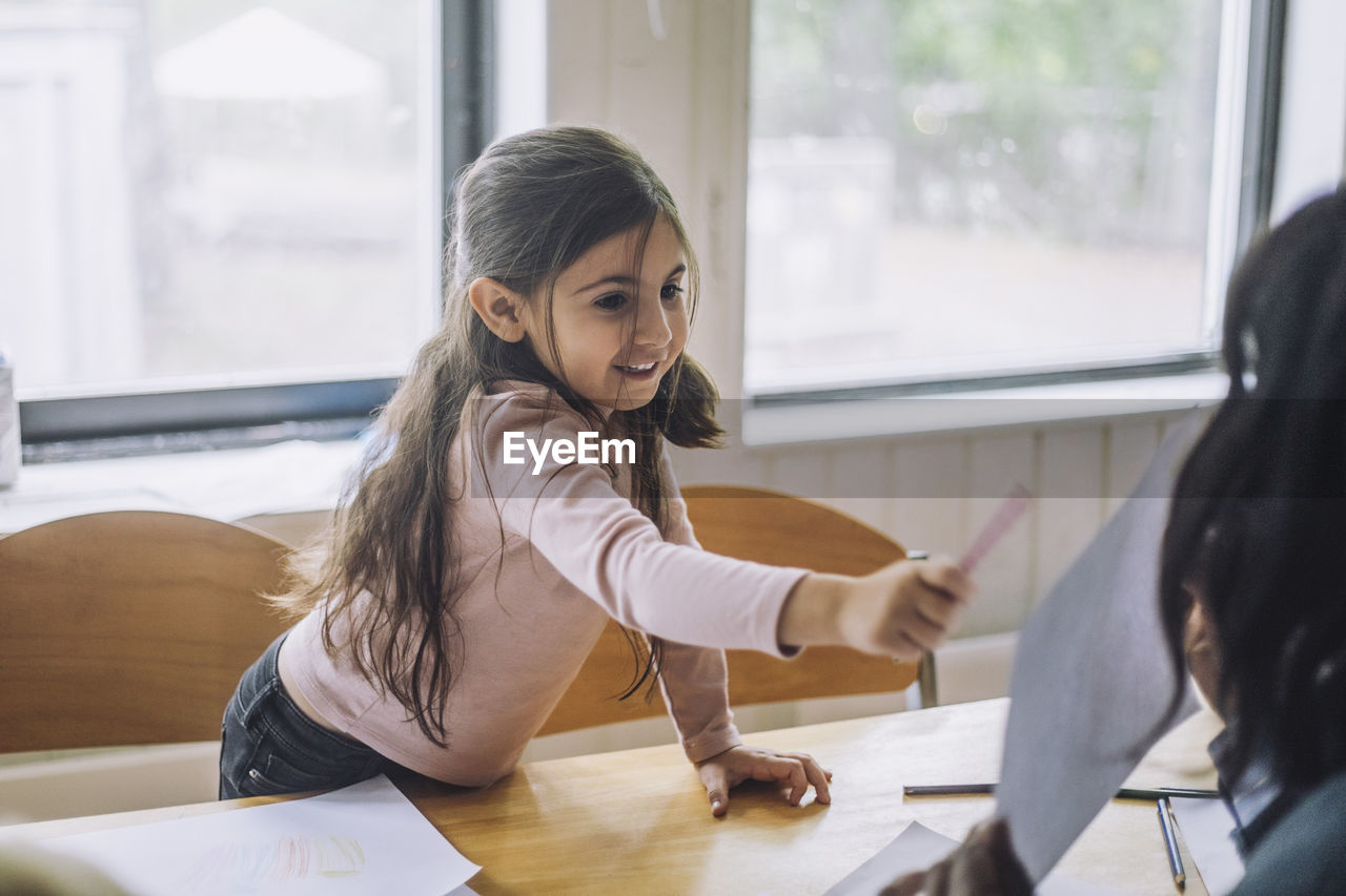 Smiling girl pointing at teacher showing drawing during art class in kindergarten