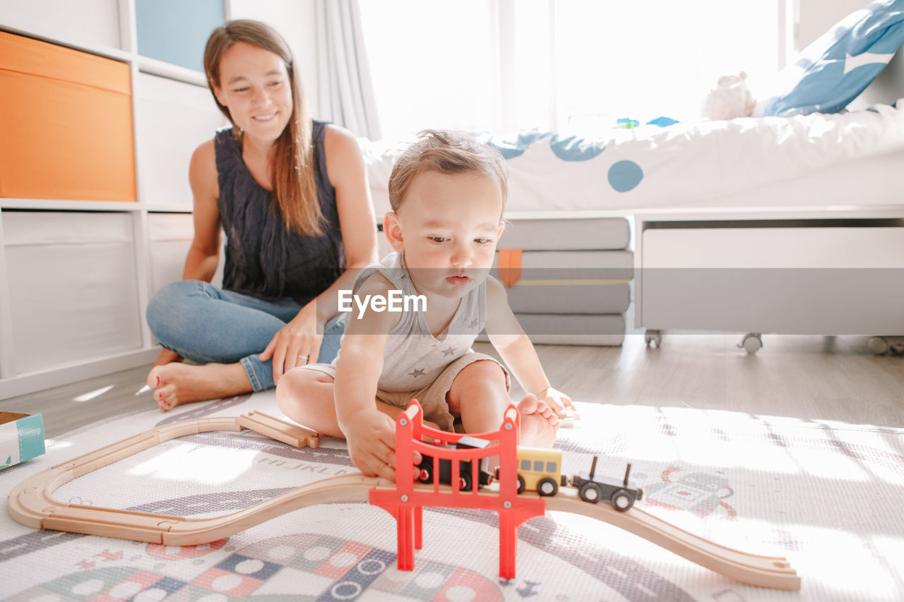 Mother and toddler boy playing with car wooden railway on floor at home. early age education 