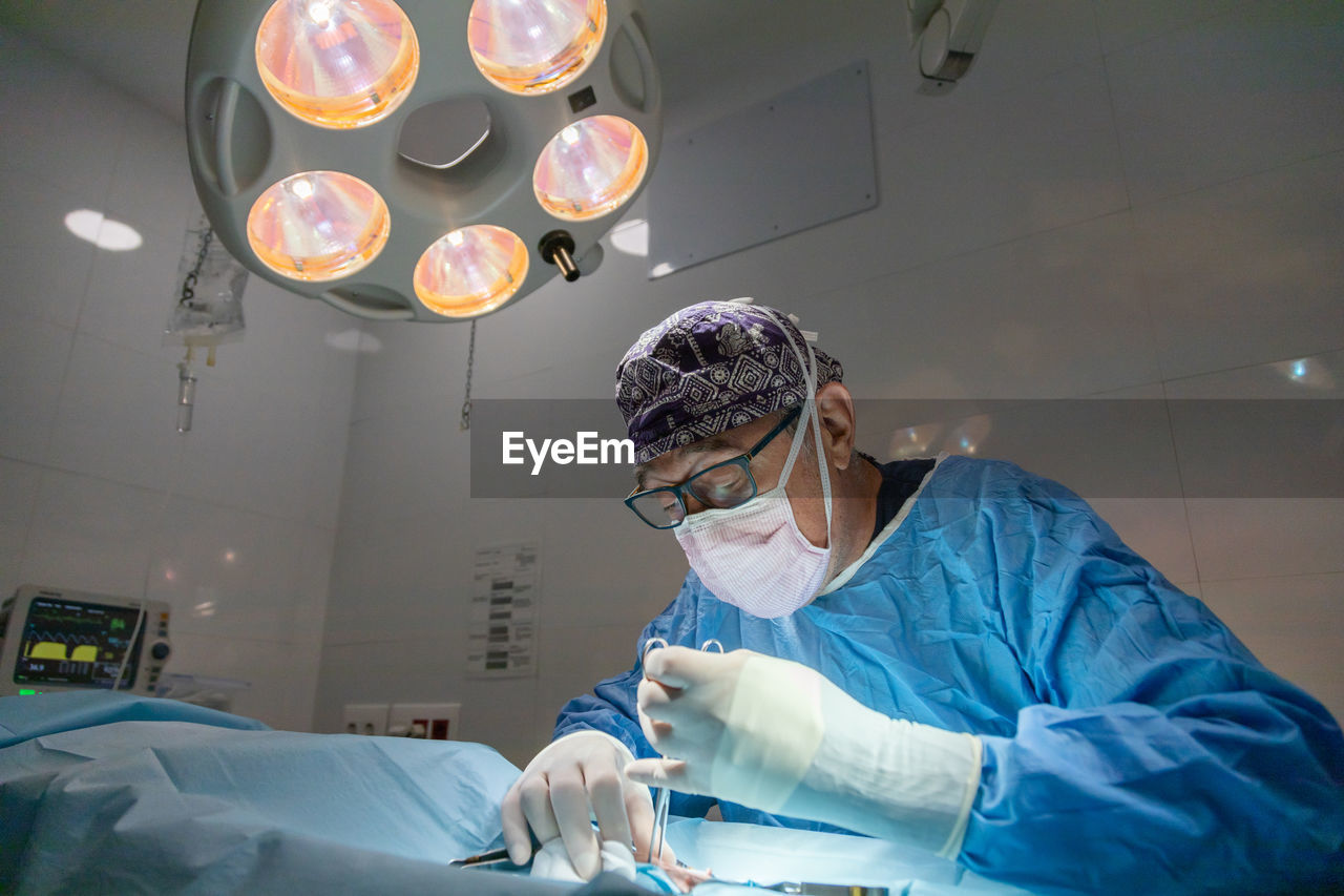 Professional senior male surgeon in mask and uniform doing operation under lamp in operating room