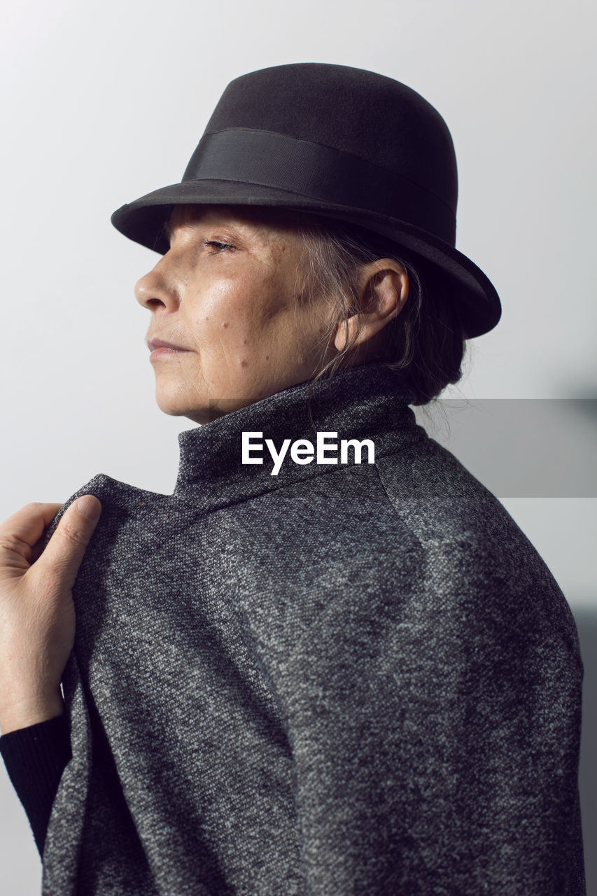 Fashionable older woman in black clothes and a hat stands against a white wall in the office