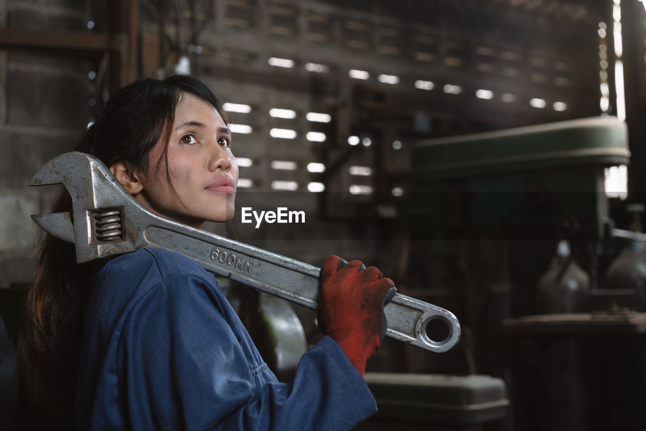 Side view of young woman with large wrench standing in factory