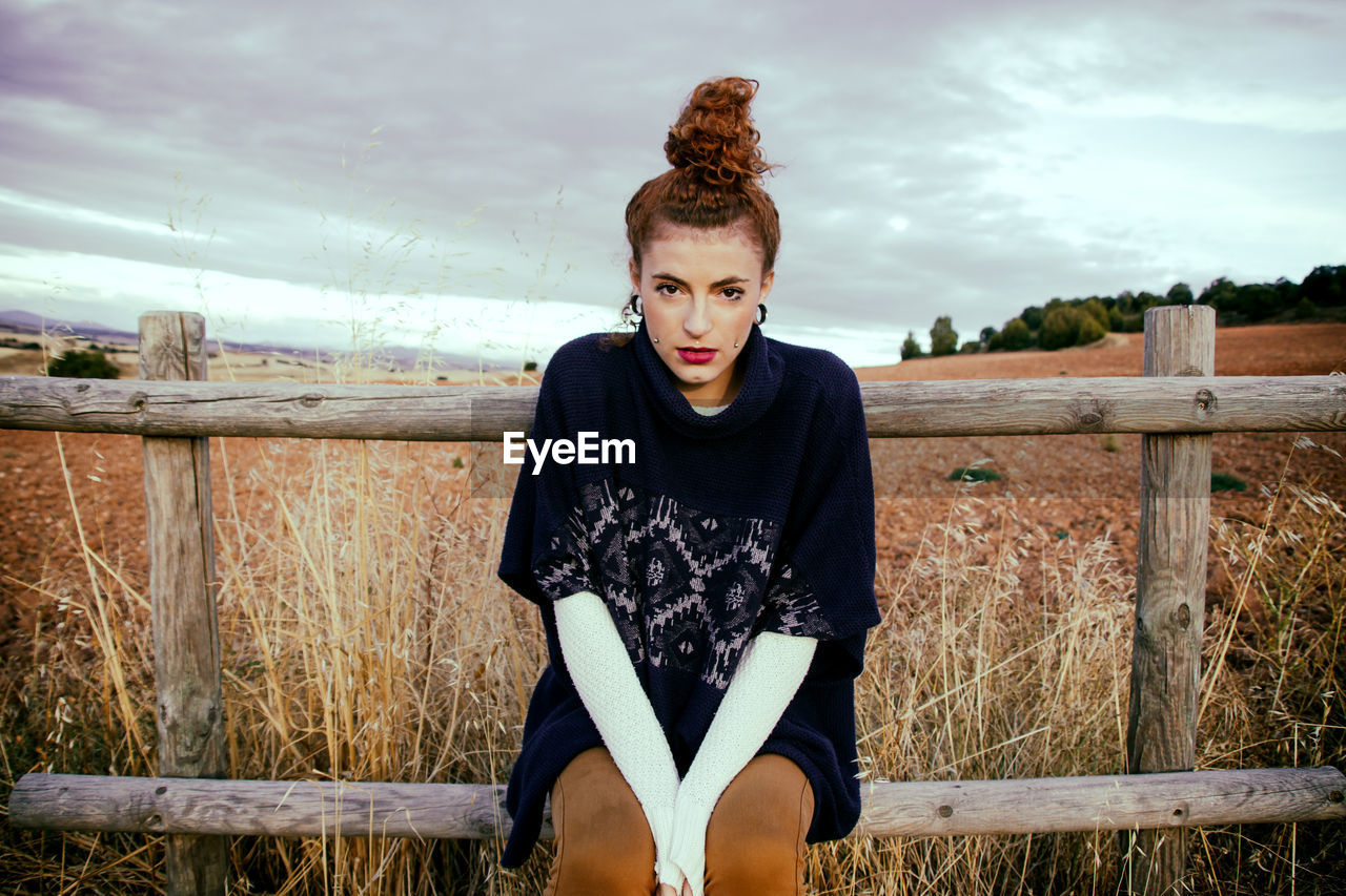 Portrait of young woman sitting on railing at field against sky