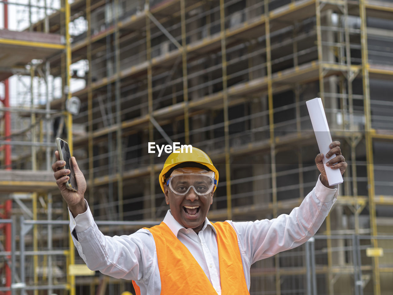 Hurrah, exulting and joyful indian engineer holding rolled up plans on construction site