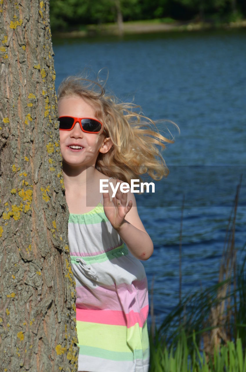 Portrait of cute smiling girl wearing sunglasses while standing by tree trunk against lake