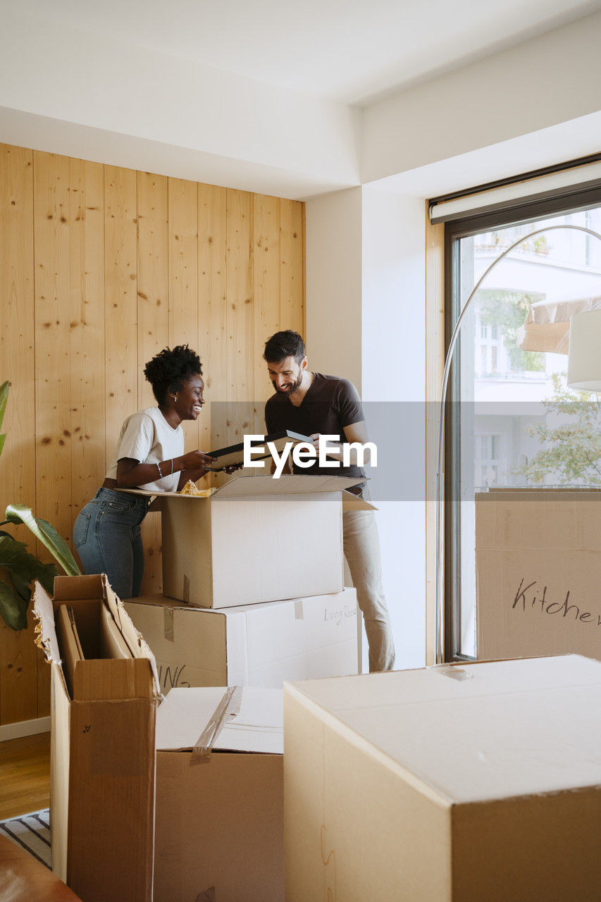 Multiracial couple removing frame from cardboard box while standing at new home