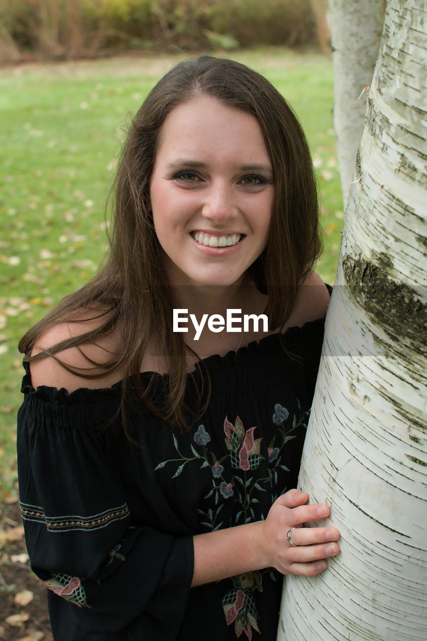 Portrait of smiling young woman standing by tree trunk