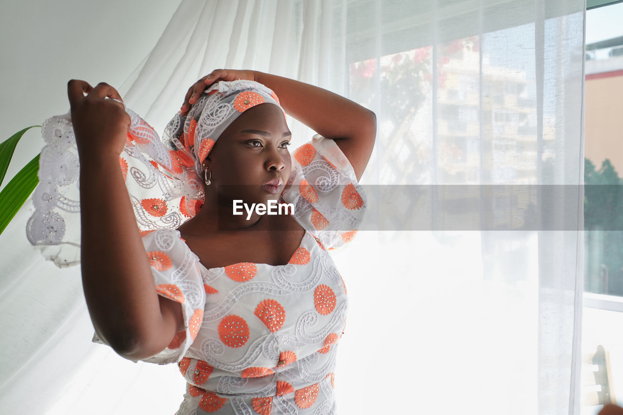 Concentrated young african lady in stylish summer dress adjusting traditional turban while standing near window in light room