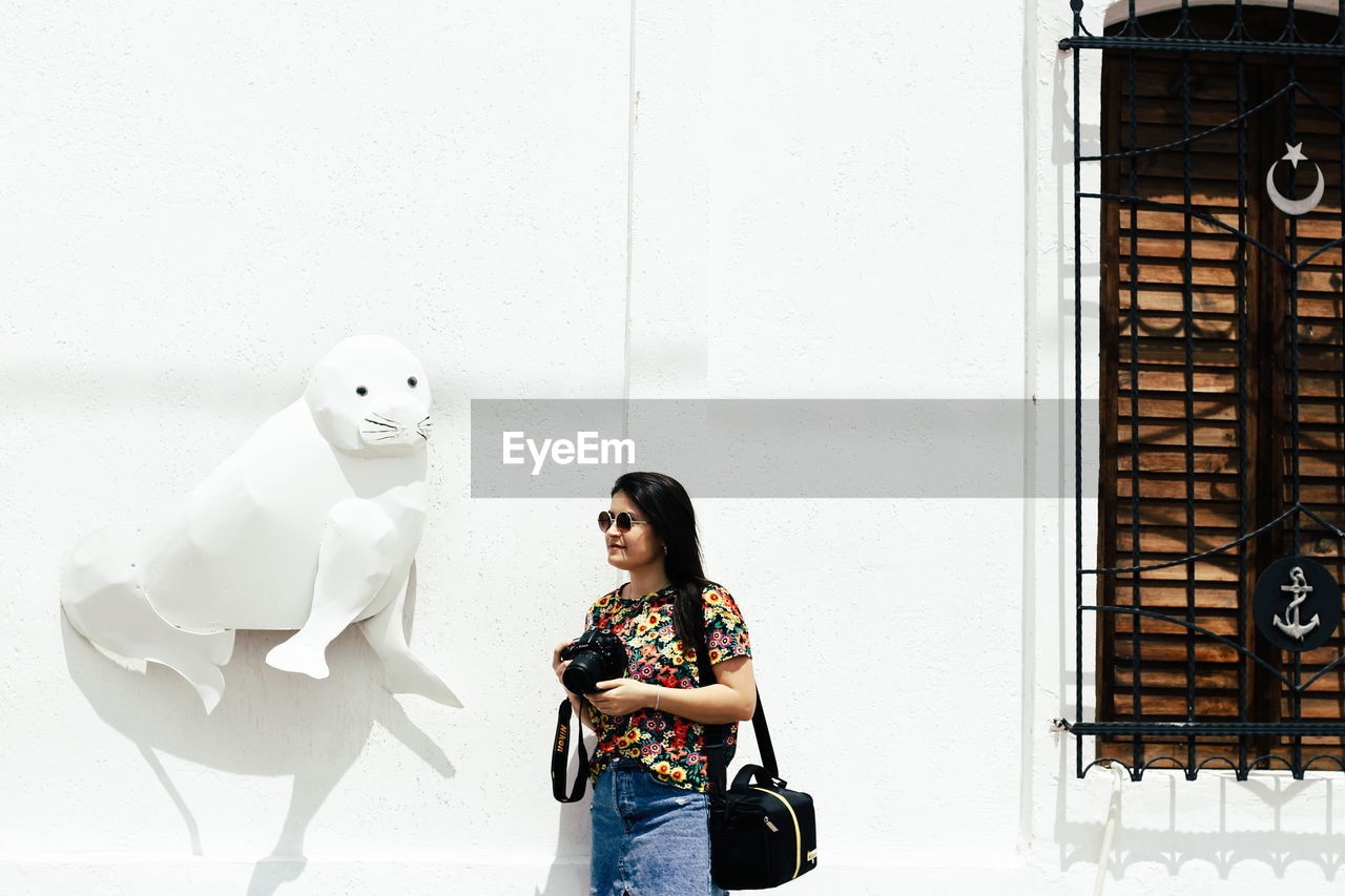 Woman holding camera while standing by wall outdoors