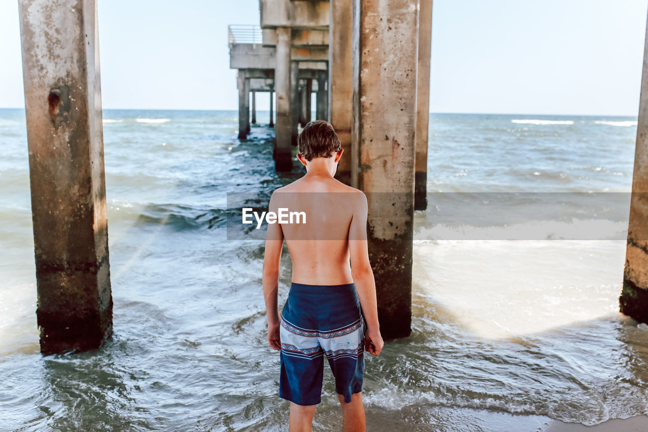 FULL LENGTH OF SHIRTLESS MAN STANDING ON WOODEN POST AT BEACH