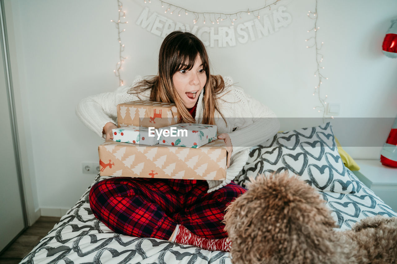 Surprised woman holding christmas presents while sitting with dog on bed