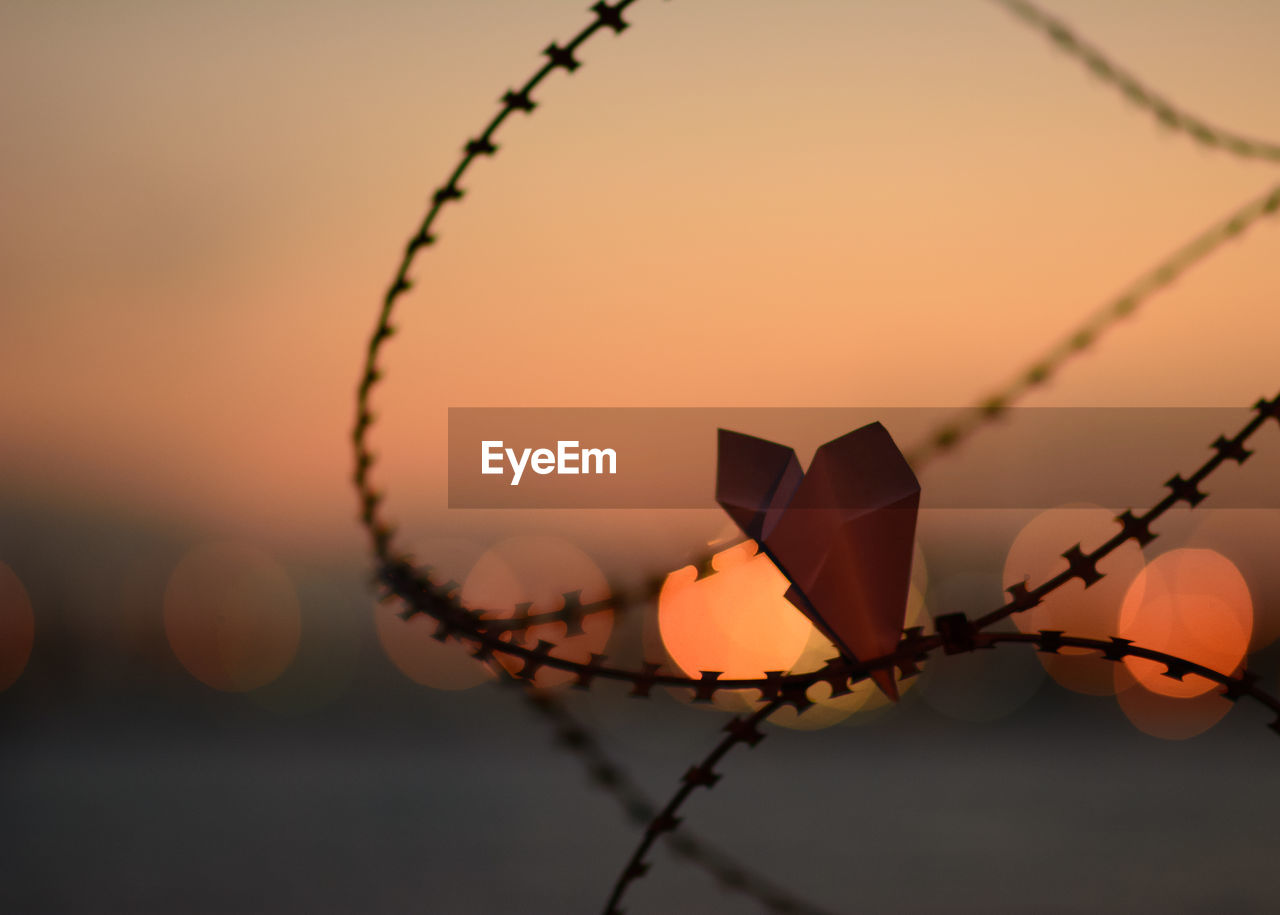 Close-up of paper airplane in barbed wire against sky during sunset