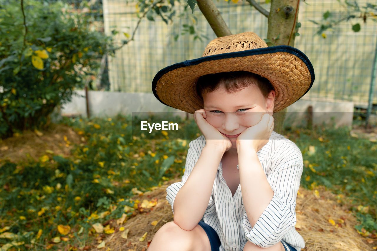 Cpre-school student in a straw hat and a rustic white shirt sits under an apple tree in home garden