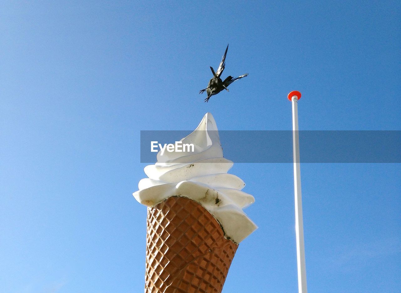 Low angle view of bird flying over ice cream against clear blue sky