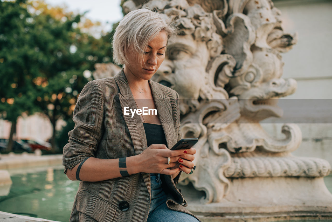 Blond woman looking at her smartphone while sitting at fountain