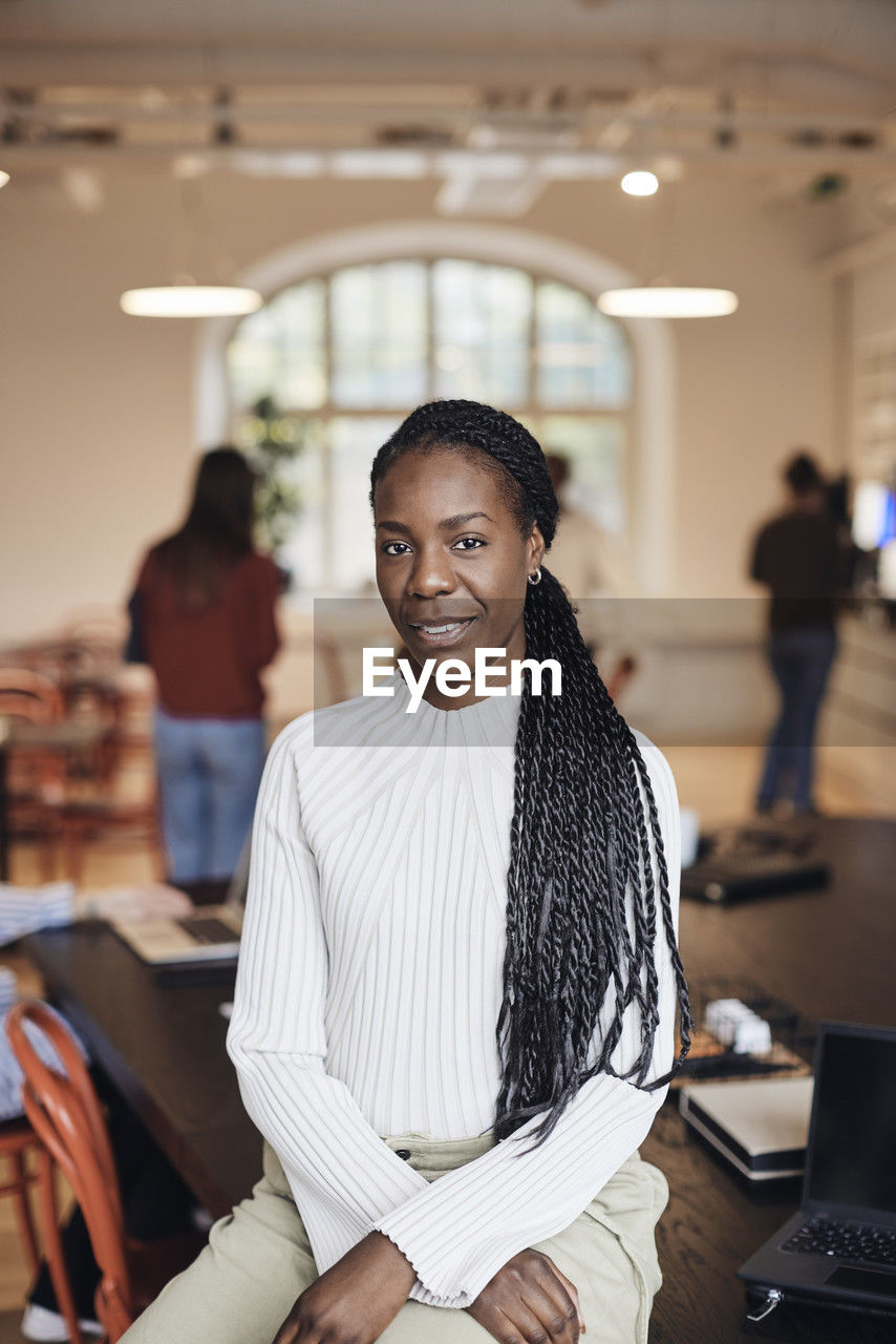 Portrait of confident female business professional with braided hair sitting at desk in office