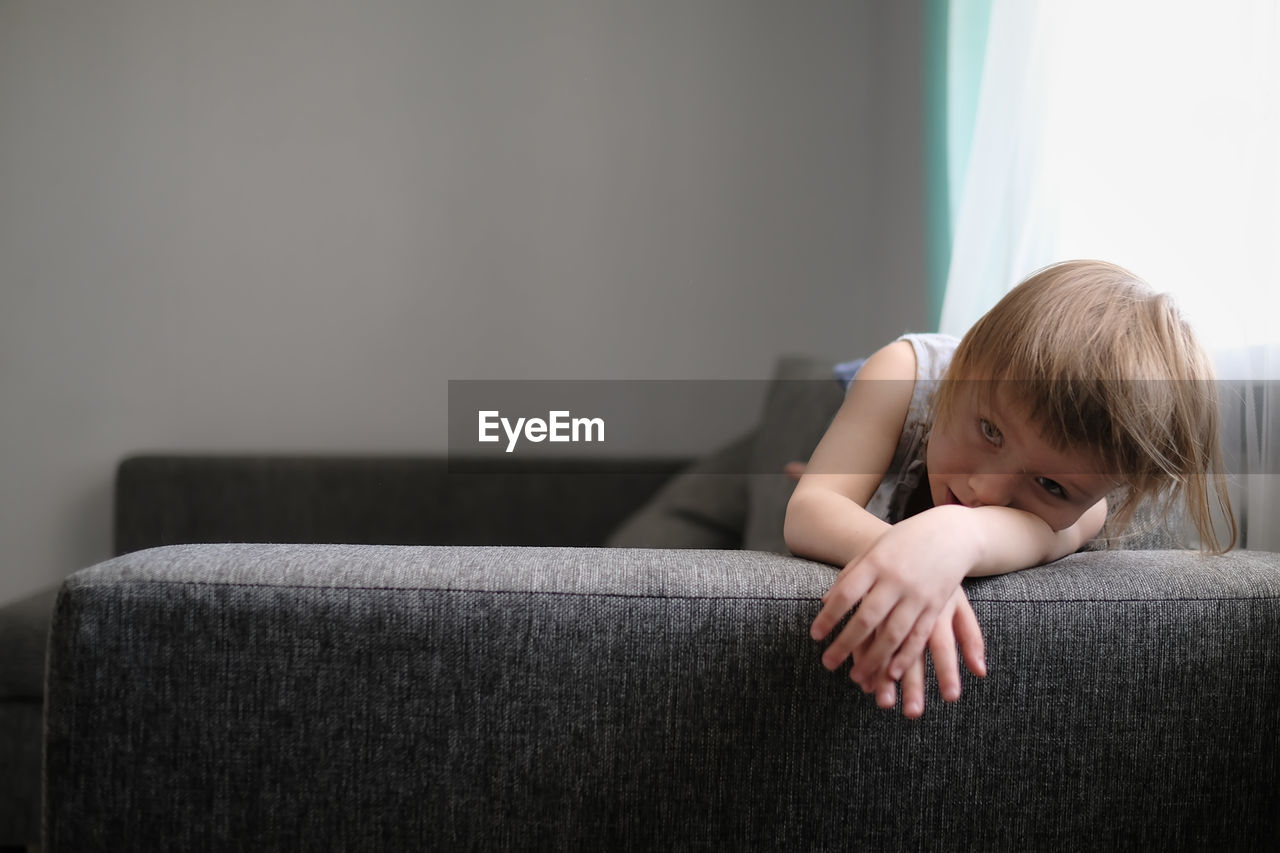 European toddler child sits and gets bored on gray sofa in bright living room, minimalist interior