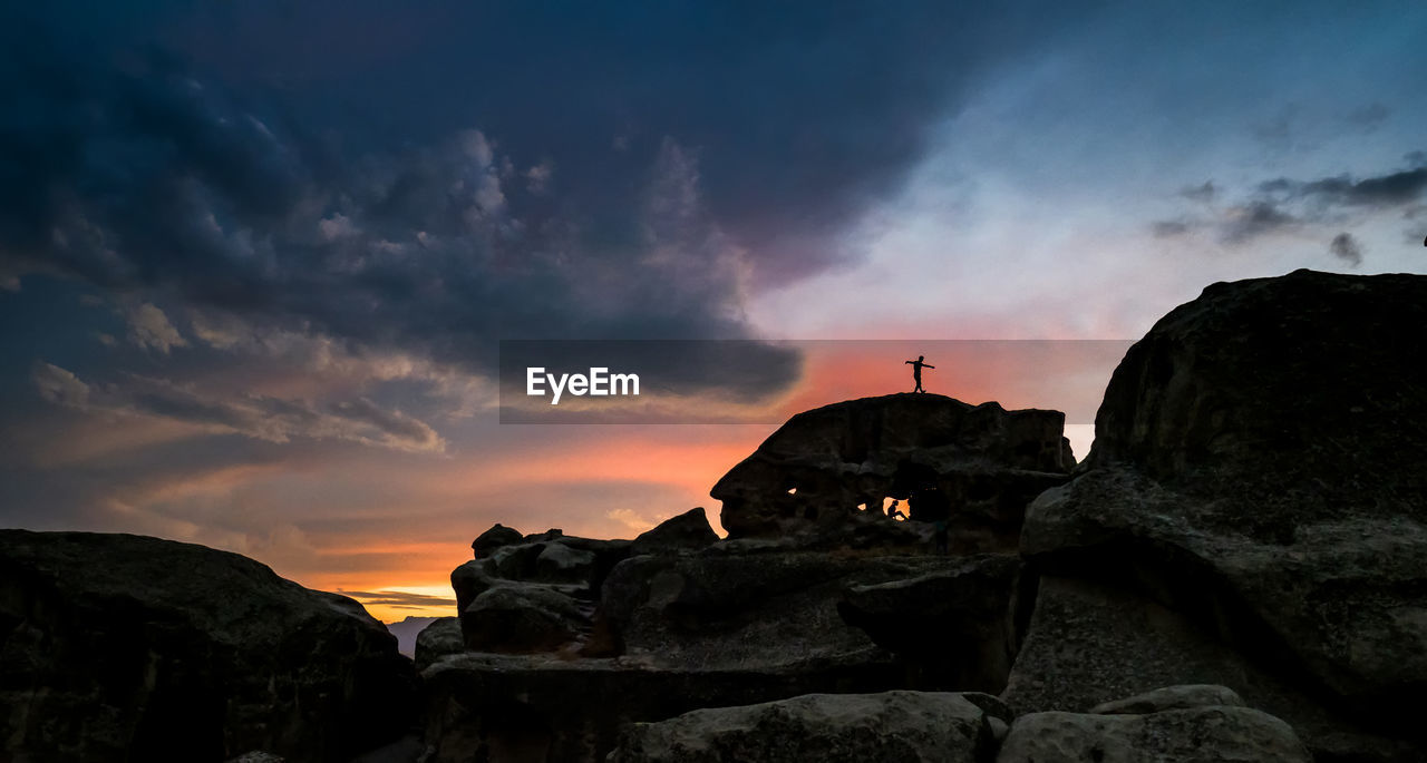 Distant view of silhouette man on rock against cloudy sky at sunset