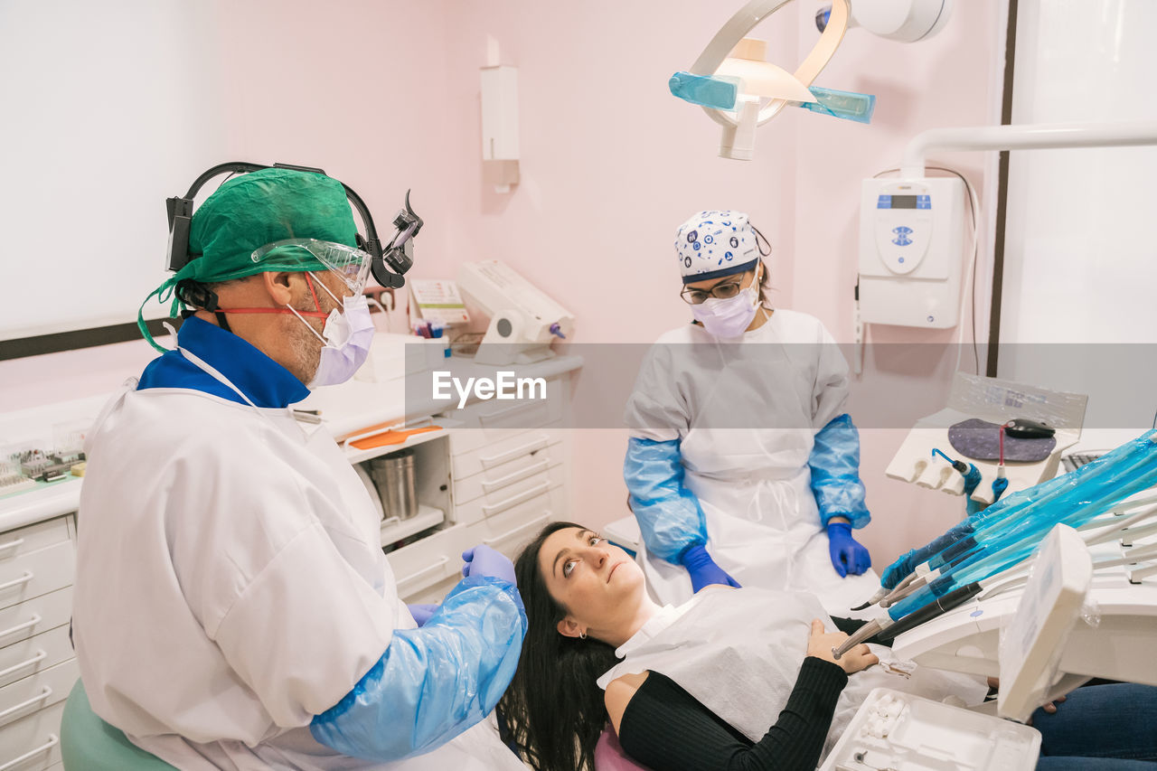 From above of anonymous stomatologist and assistant in uniforms talking to woman lying on medical chair near professional equipment