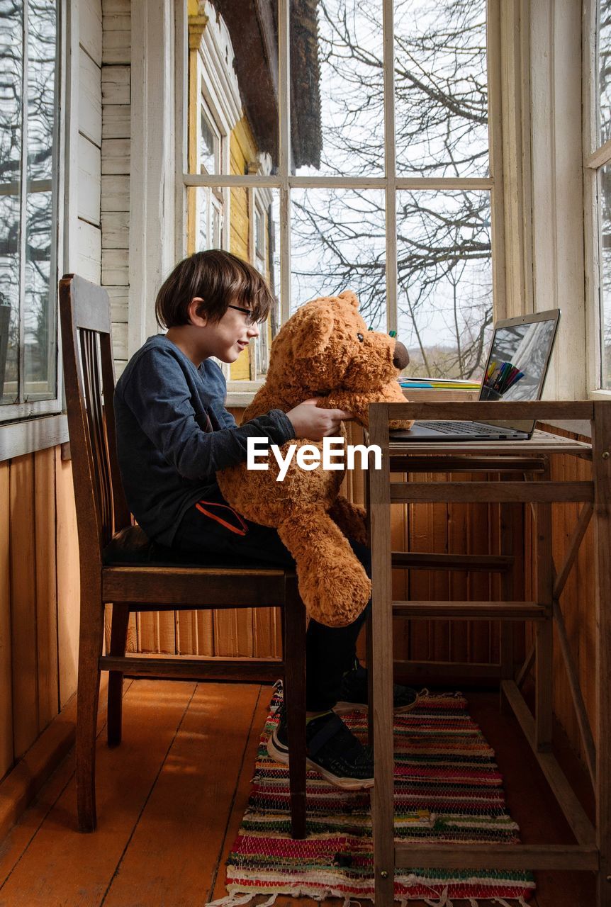 Kid sitting on chair, holding toy bear, looking at computer