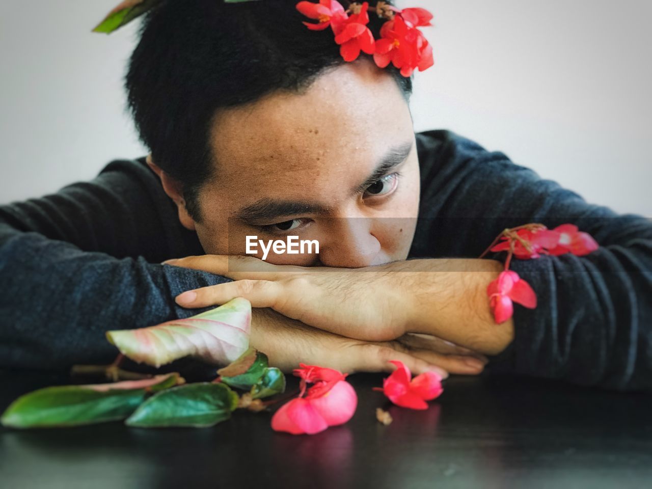 Close-up of young man with red begonia flowers resting his head on forearms on table.