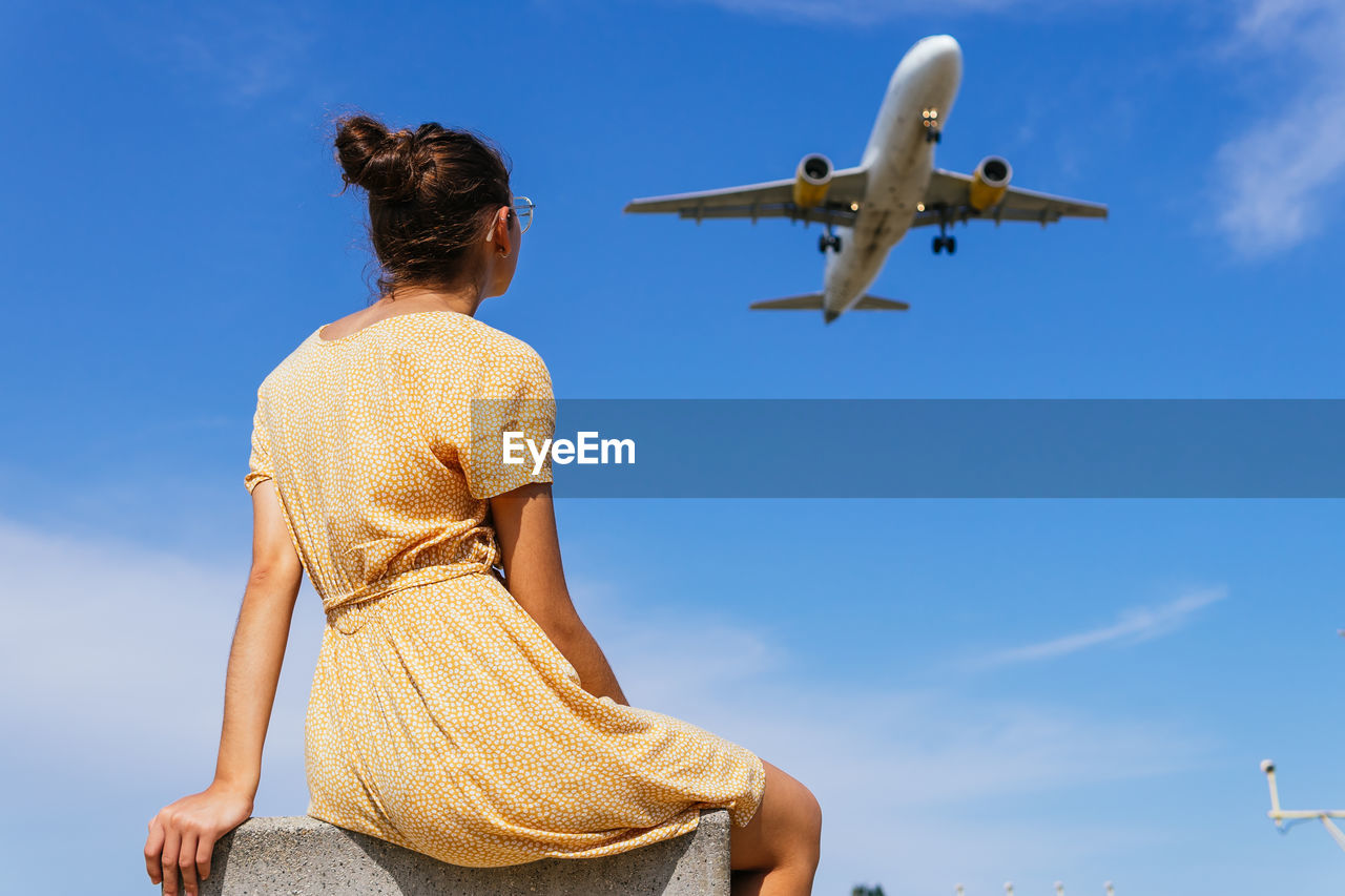 From below back view of unrecognizable female in casual clothes sitting against blue sky with plane flying in blue sky