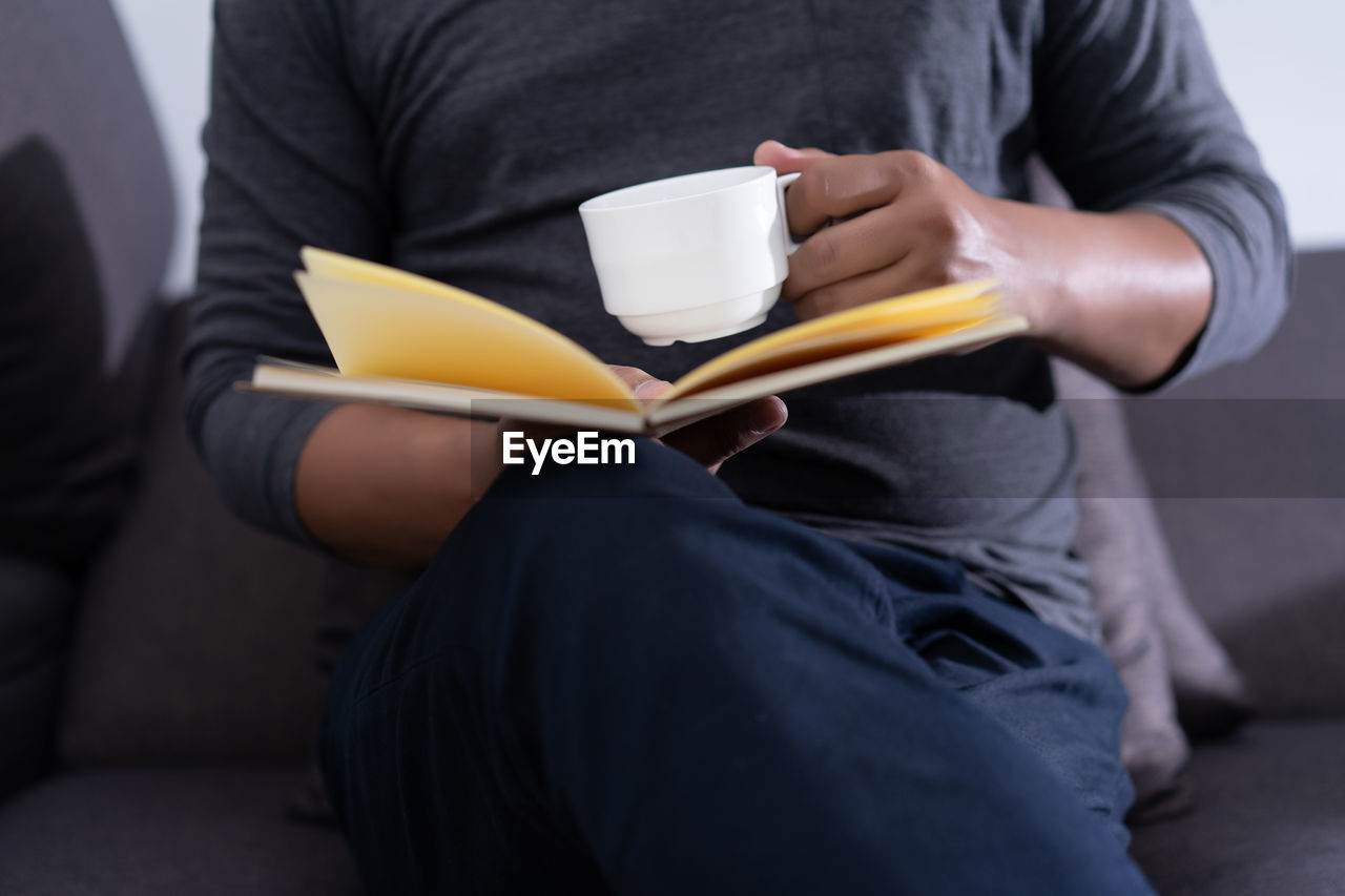 Midsection of man reading book while having drink at home