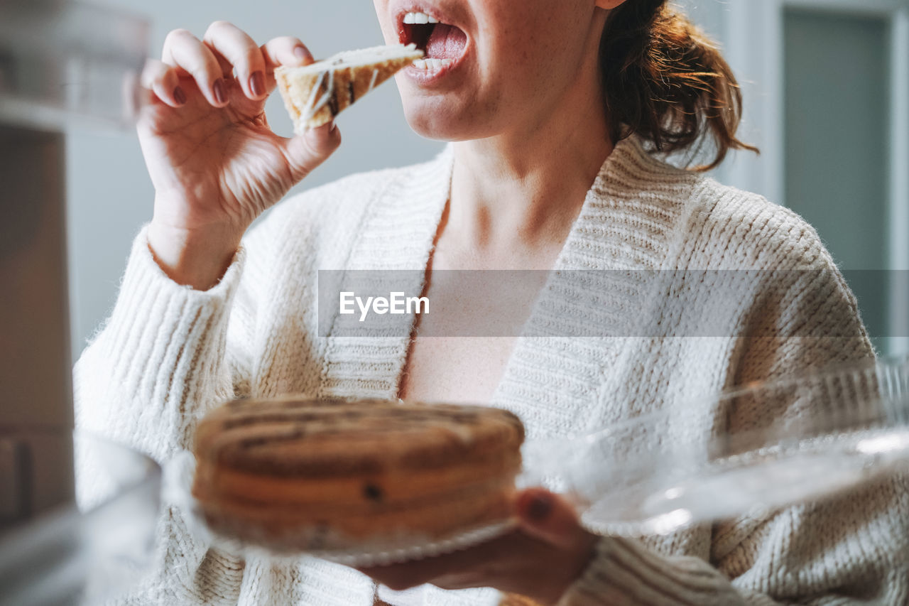 Attractive friendly brunette woman middle age in cozy cardigan eating cake from refrigerator 