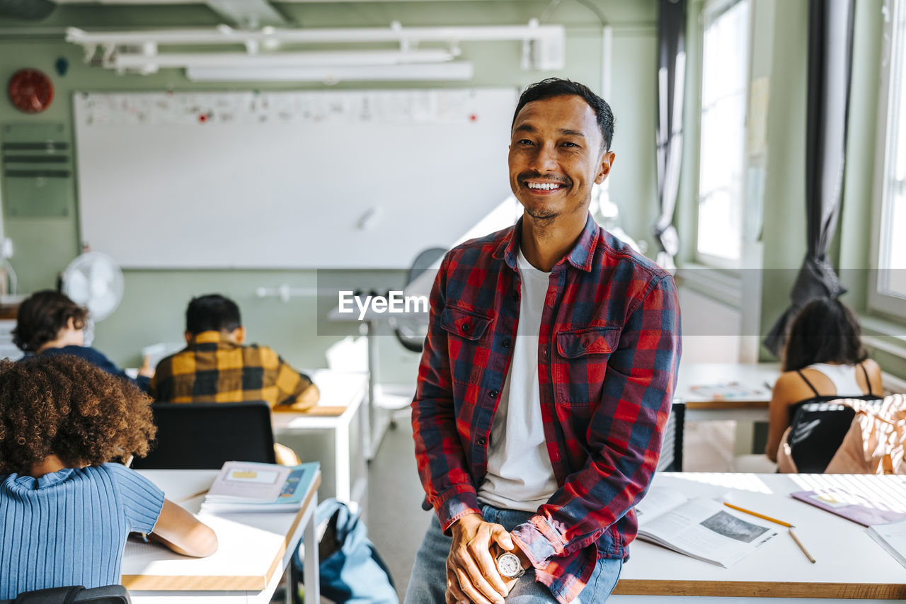 Portrait of happy male teacher in plaid shirt sitting on desk in classroom at elementary school