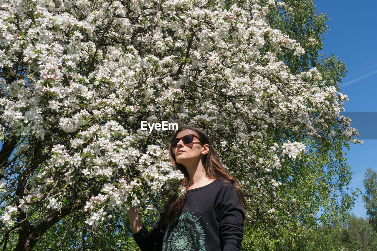 PORTRAIT OF BEAUTIFUL YOUNG WOMAN STANDING BY FLOWER TREES