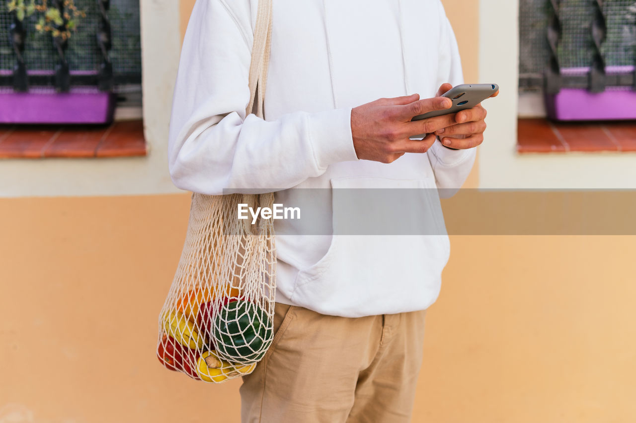 Cropped unrecognizable male standing on city street with eco friendly mesh bag with groceries and using smartphone