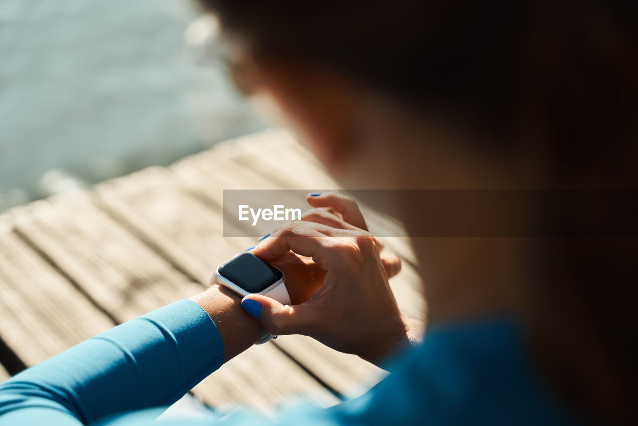 From above back view of unrecognizable female athlete sitting on mat and checking pulse on fitness tracker while resting after outdoor workout