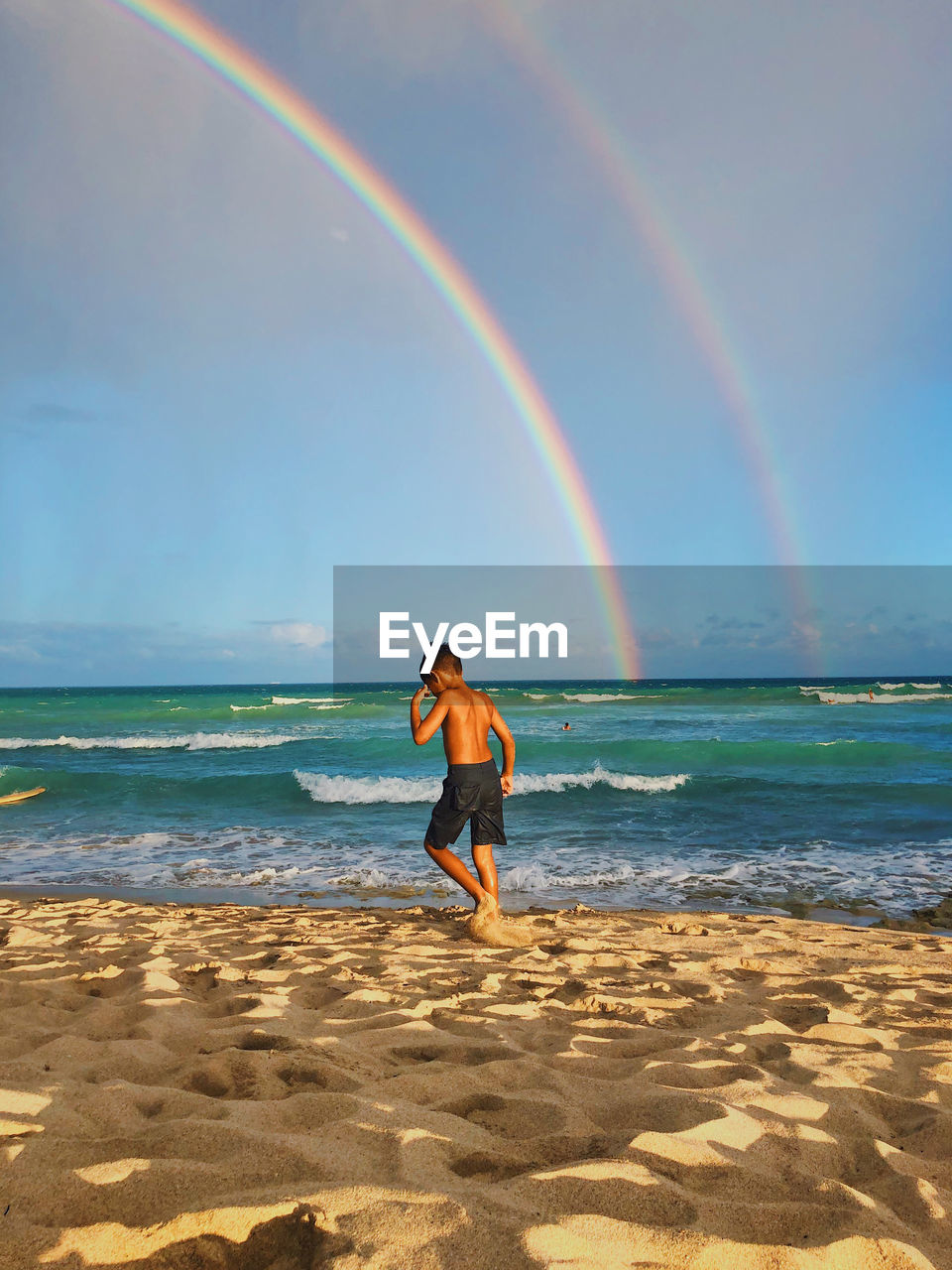 Shirtless boy walking against rainbow at beach