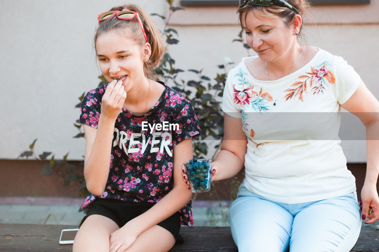 Smiling mature woman holding blueberries in glass by daughter sitting on bench