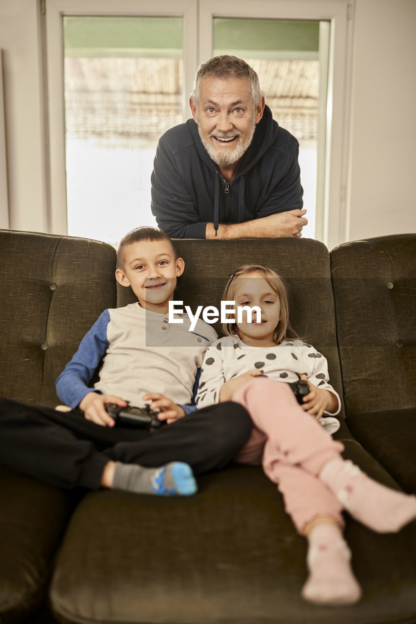 Girl and boy playing video game with grandfather leaning on sofa at home