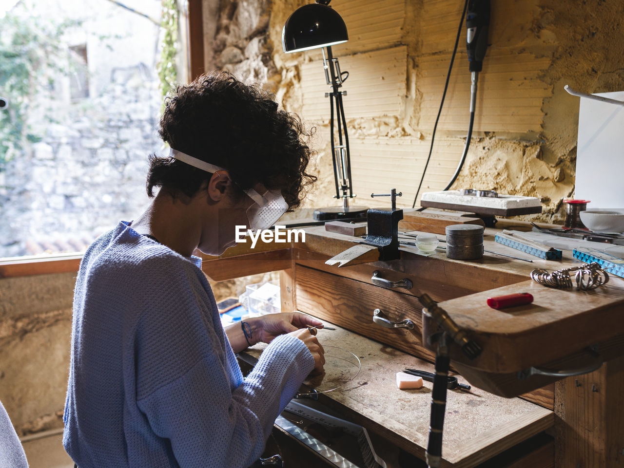 Side view of curly haired ethnic female worker in transparent glasses concentrating at goldsmith workbench working with wire