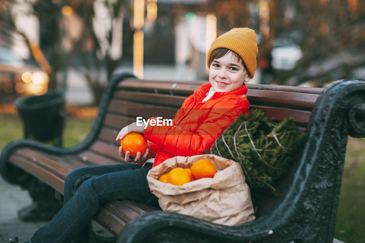 A boy in a bright orange jacket and a fashionable yellow hat sits on a bench, laughs, throws oranges