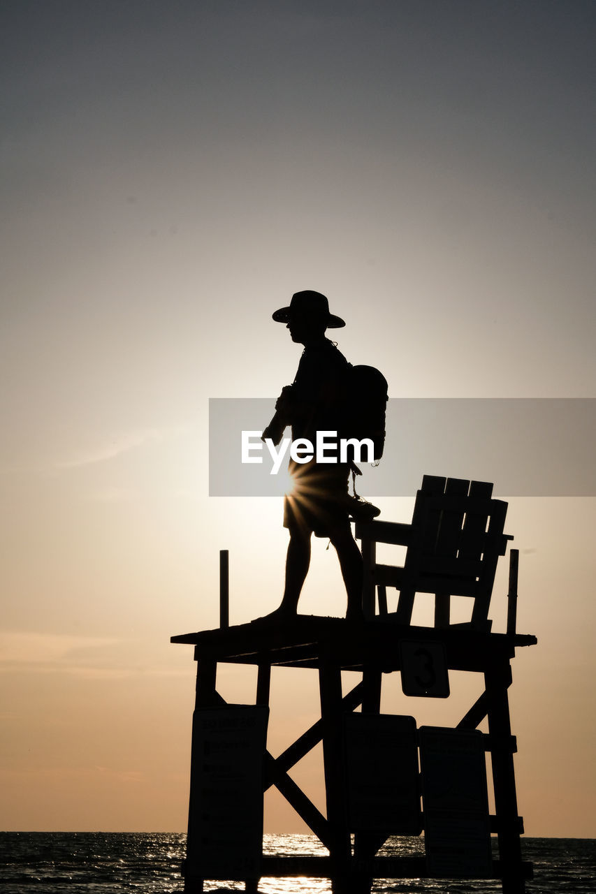 Silhouette man standing on lifeguard hut at beach against sky during sunset