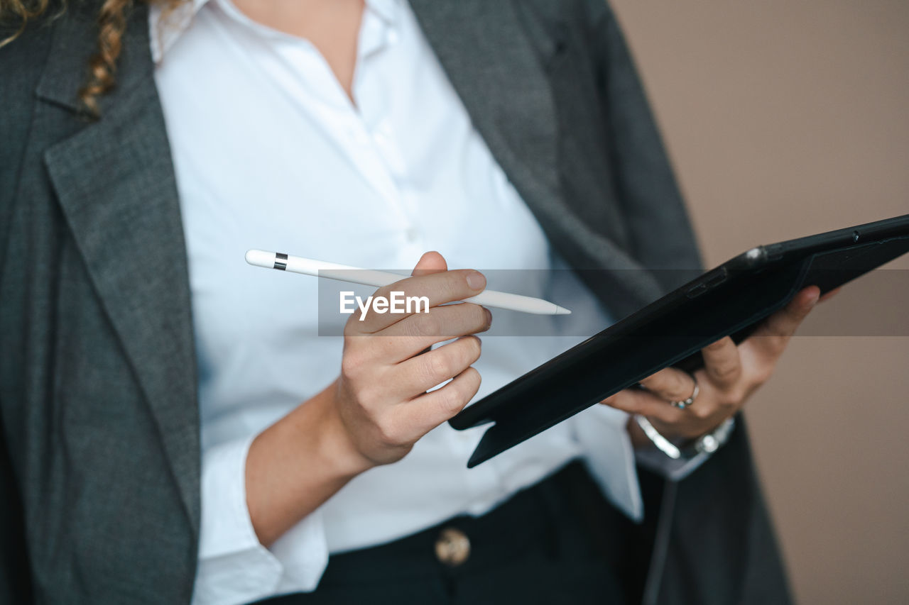 Close up of anonymous female entrepreneur in formal outfit using tablet with stylus and rejoicing over project accomplishment on brown background in studio